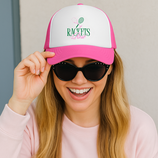 girl wearing a pink and white trucker cap with a rackets and rose design and sunglasses, smiling.