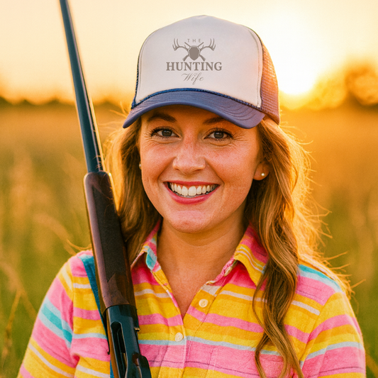 Woman wearing a cap with 'Hunting Wife' text, holding a rifle in a field at sunset.