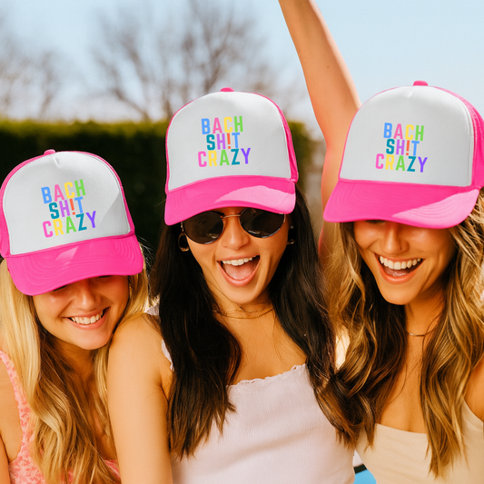 Three women wearing pink and white hats with colorful text that says bach sh!t crazy outdoors.
