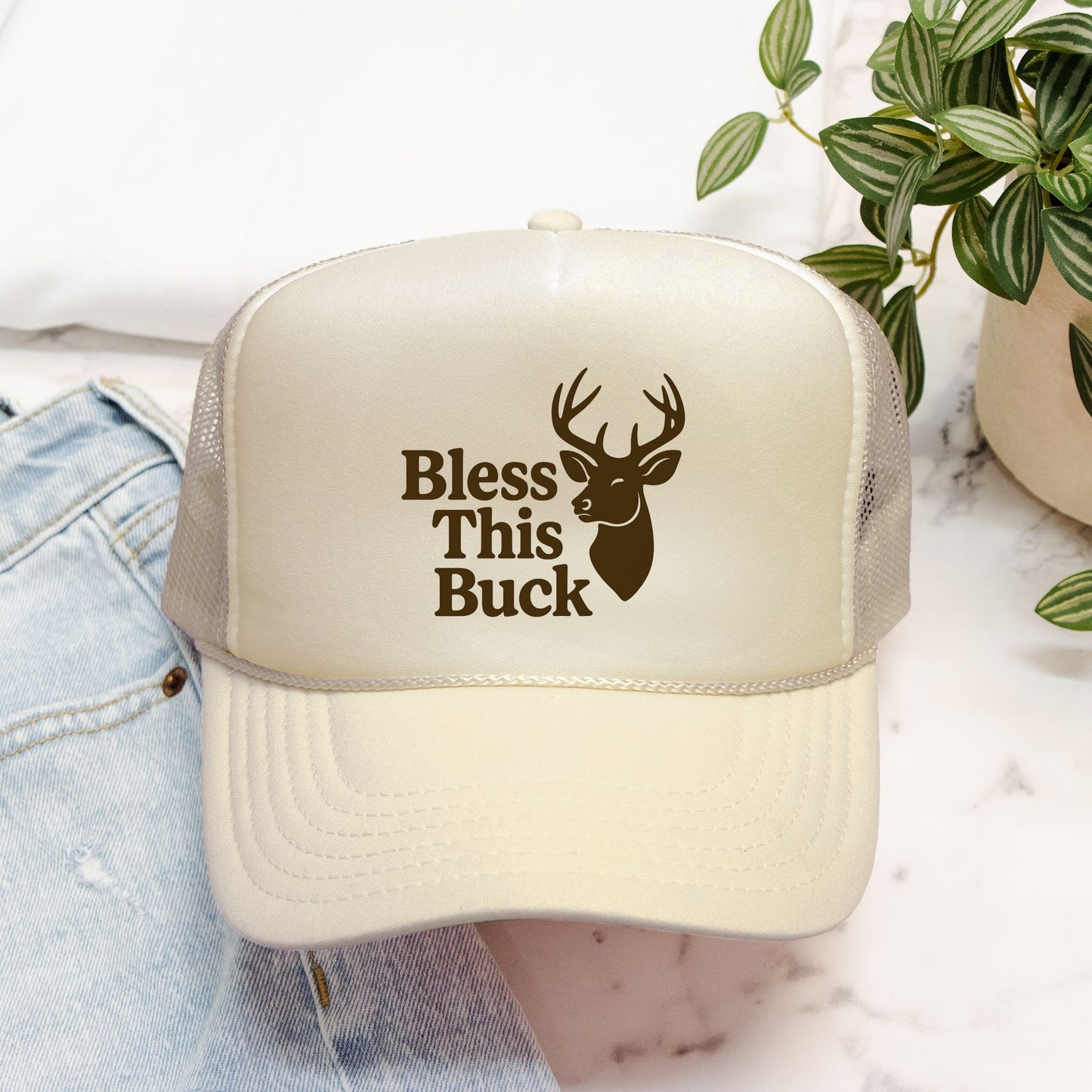 Beige trucker hat with 'Bless This Buck' text and deer graphic on a white surface.
