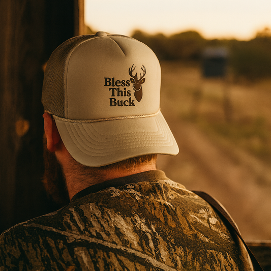 Man wearing a cap with 'Bless This Buck' text and a deer graphic, standing on a dirt road at sunset.