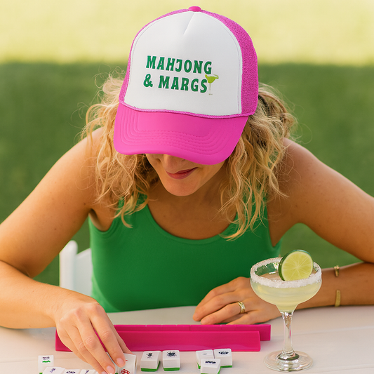 Woman wearing a 'Mahjong & Marg' cap playing mahjong with a margarita on a table.