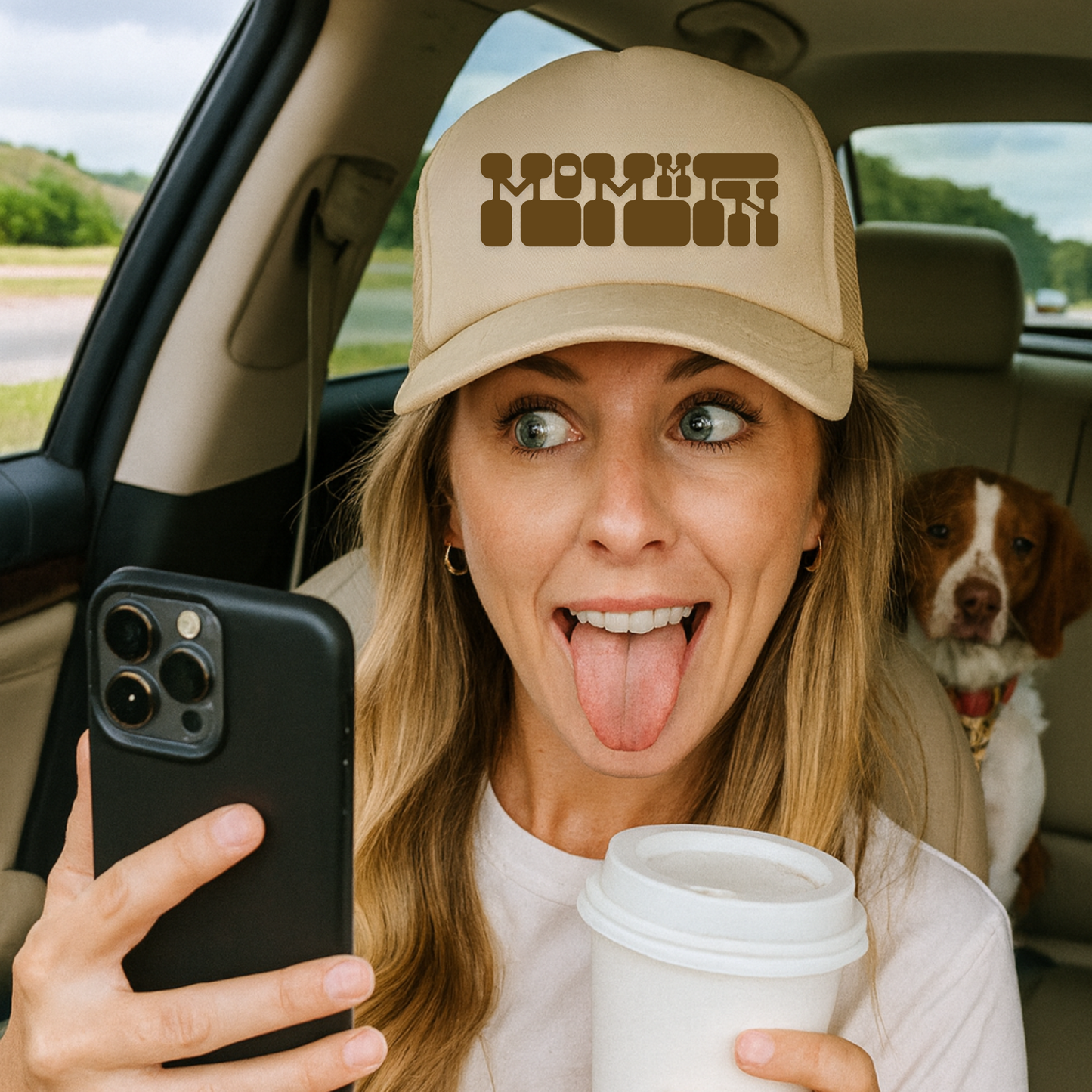 Woman in a car with a dog, holding a phone and a cup, wearing a cap with text.