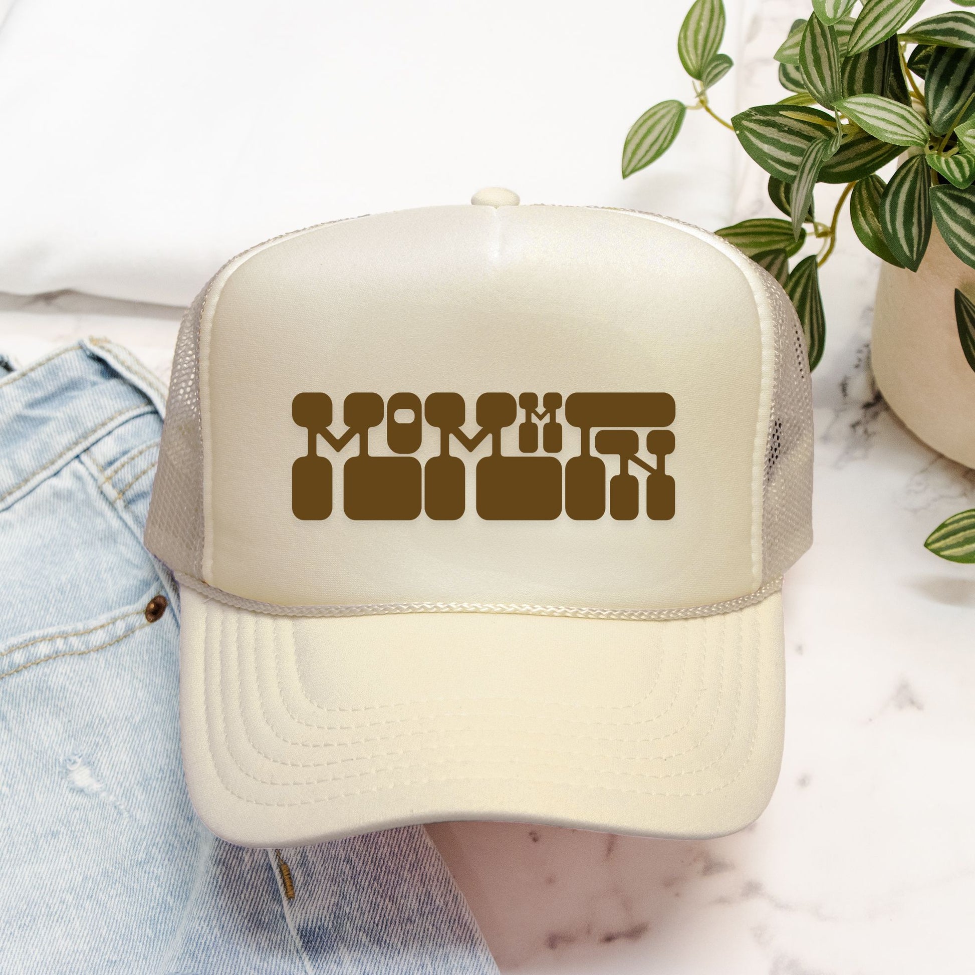 Beige trucker hat with brown text on a white surface with a plant in the background