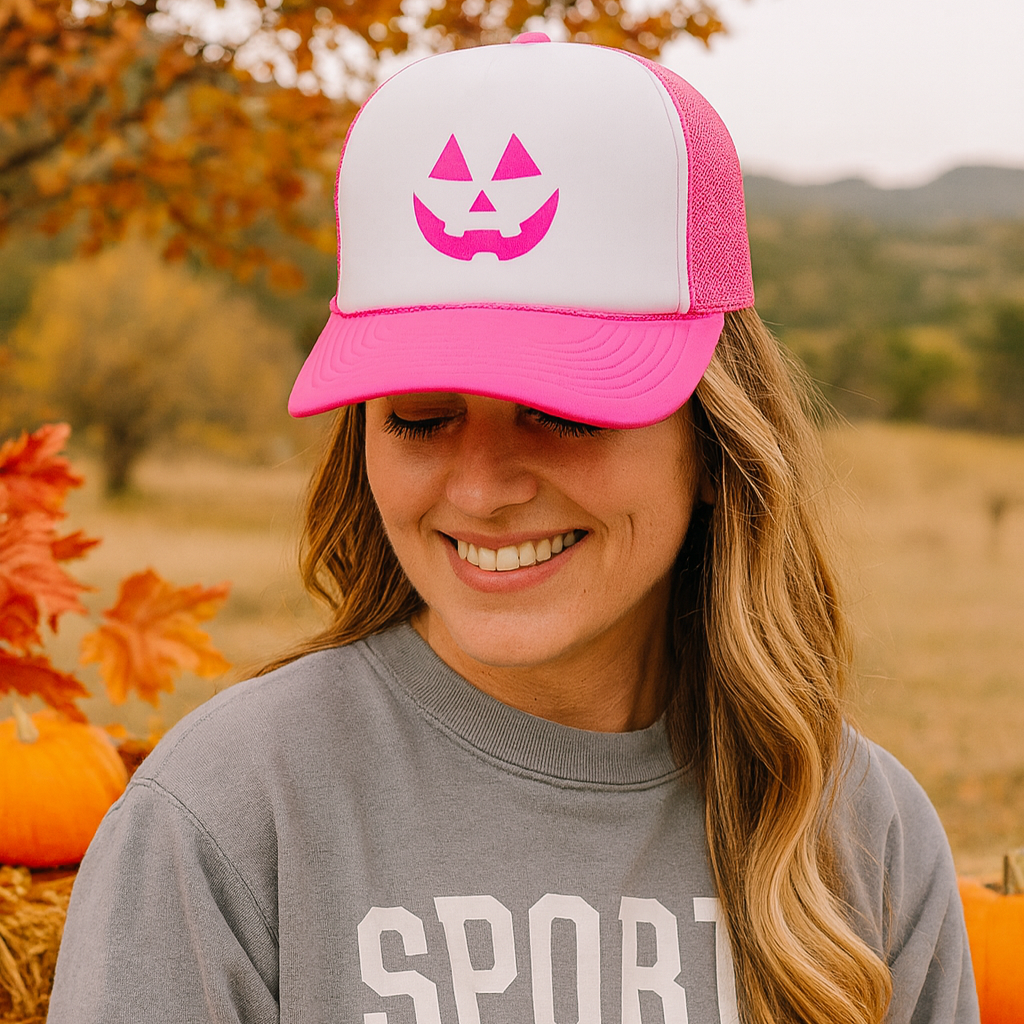 Woman wearing a pink and white cap with a pumpkin face design, smiling in a field with pumpkins and trees.